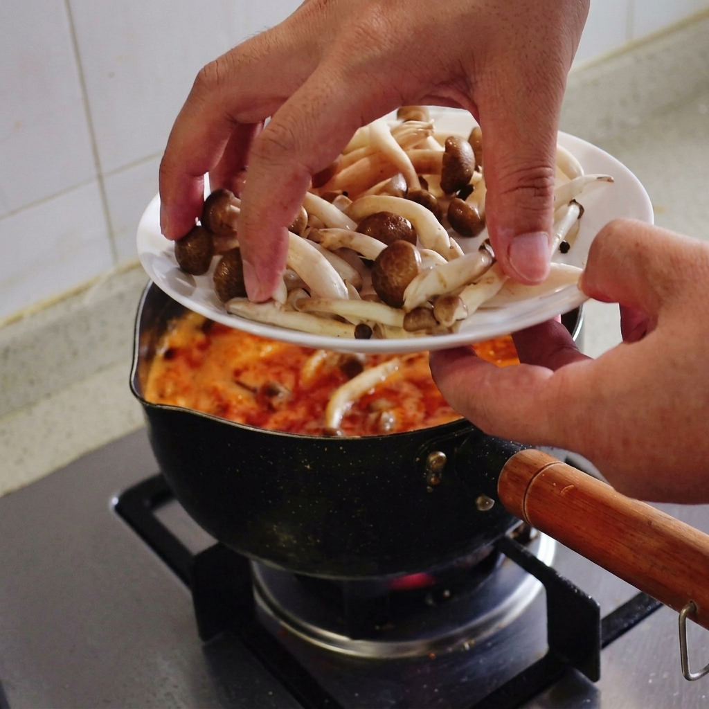 沸騰する赤いスープの鍋に、白いお皿から新鮮なしめじを手で入れている様子。