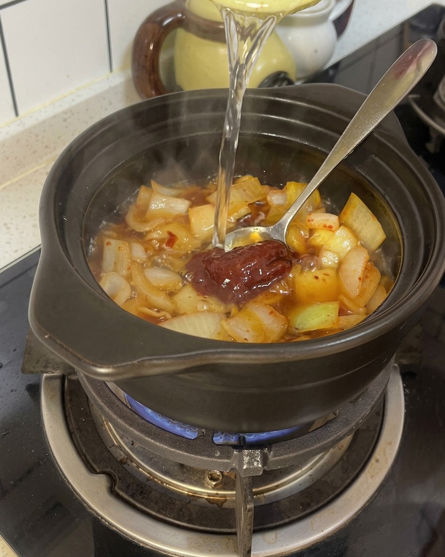 味付けした玉ねぎとコチュジャンが入った黒い土鍋に水を注いでいる様子。