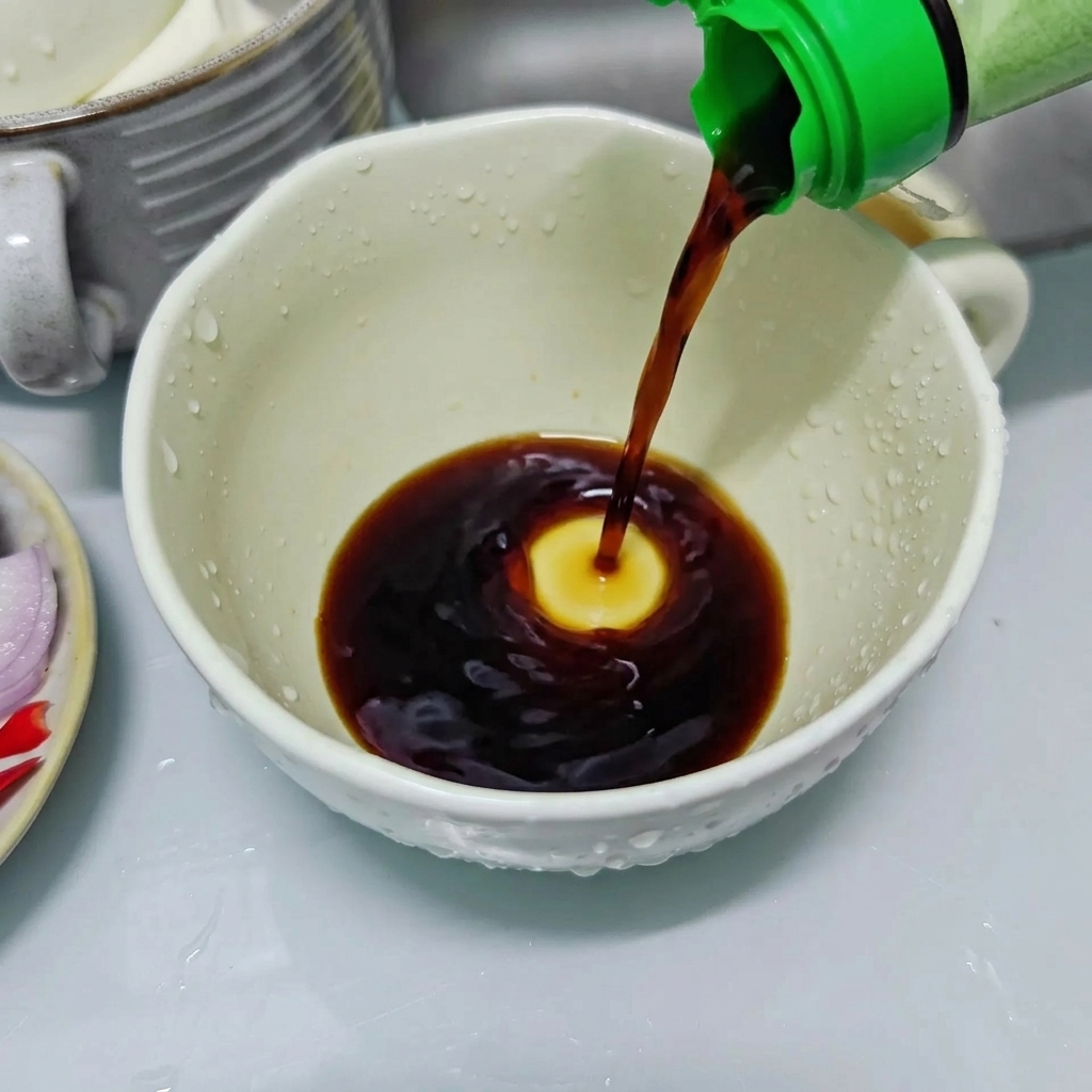味付け用のソースを準備するため、緑色のキャップのボトルから白いセラミックカップに薄口醤油を注いでいる様子。