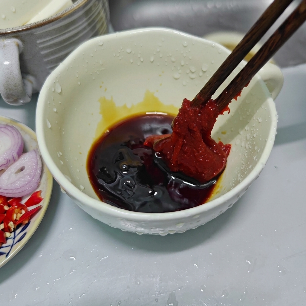 濃い色合いの合わせ調味料が入った白いセラミックボウルの上で、木箸でたっぷりの濃厚な赤色のコチュジャンを持ち上げている様子。