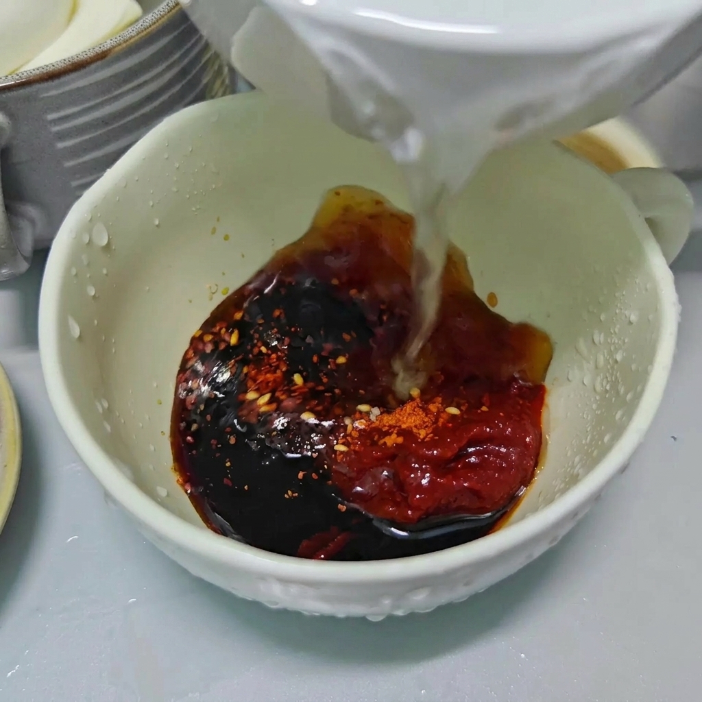 薄口醤油、コチュジャン、粉唐辛子が入った白いボウルに、透明な水を注ぎ入れている様子。