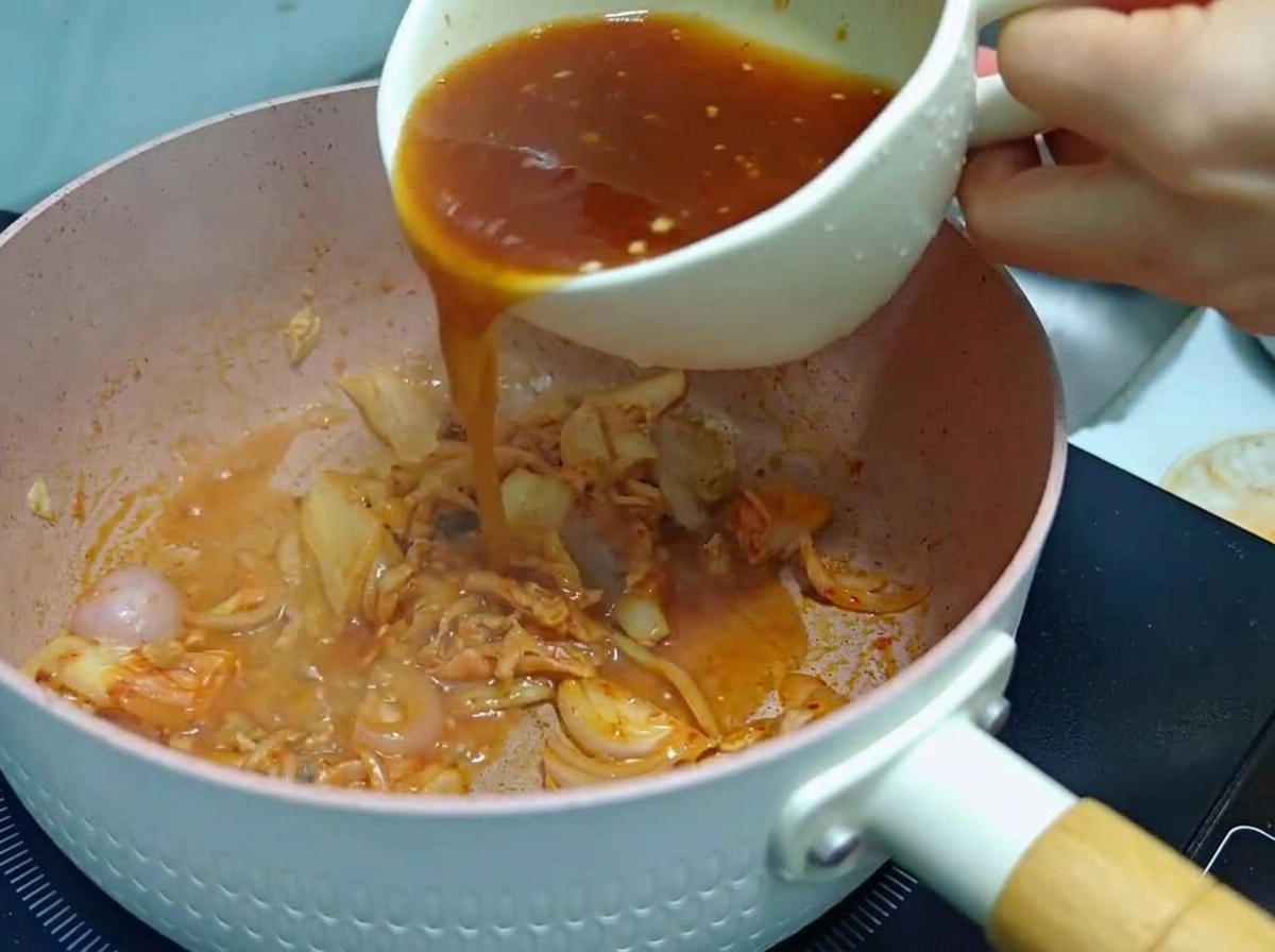 炒めたキムチと玉ねぎが入ったピンク色の調理鍋に、白いマグカップから赤茶色の合わせ調味料を注ぎ入れている様子。