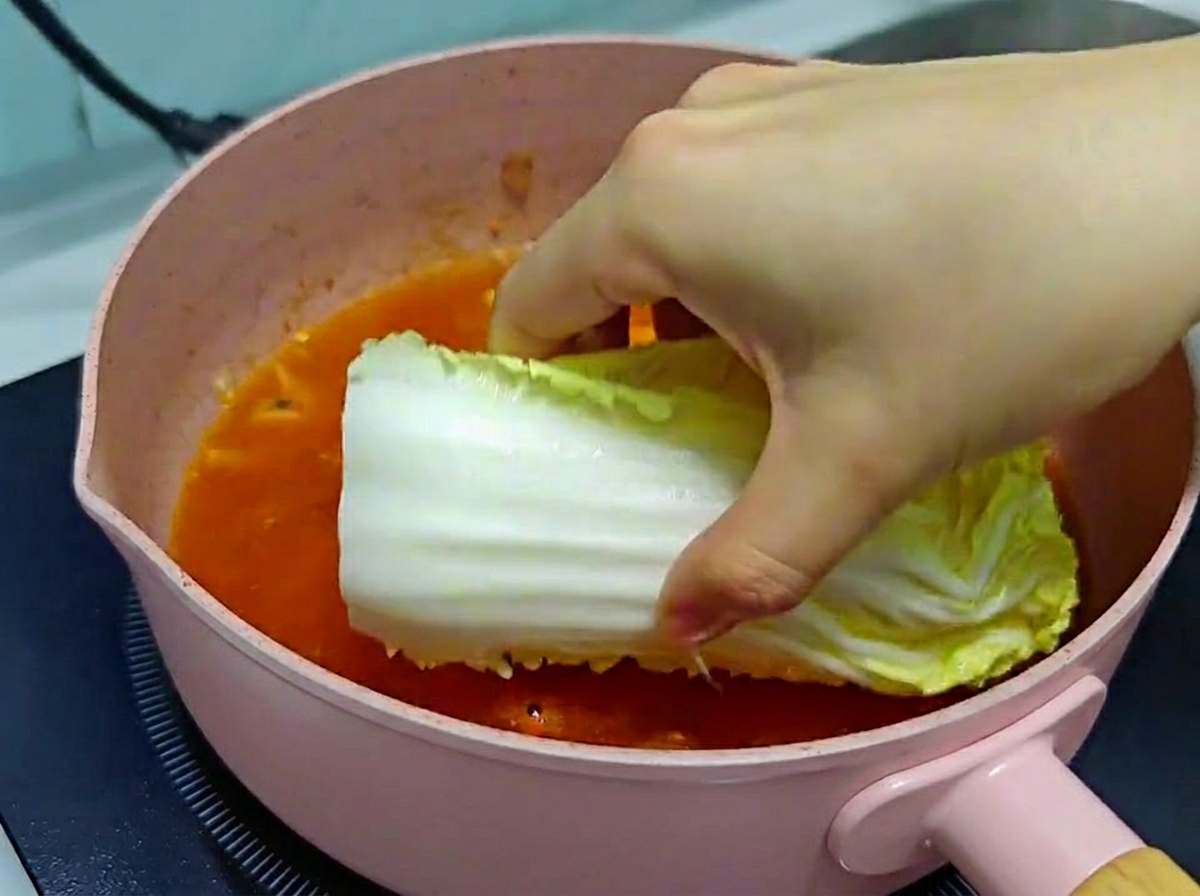 沸騰した赤いキムチスープの鍋に、新鮮な白菜の葉を丁寧に並べ入れている手元。