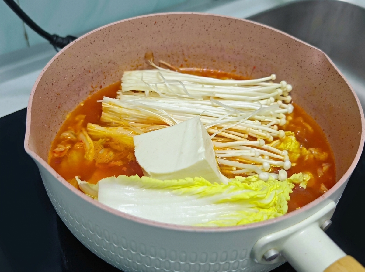 オレンジ色のキムチスープの鍋の中で、えのき茸と白菜の上にのせられた新鮮な白い大きな豆腐のブロック。