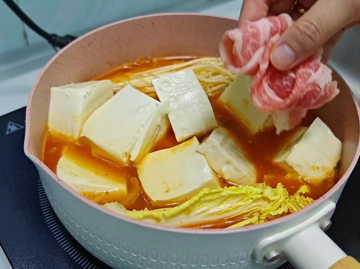 煮立つキムチスープの鍋の中で、豆腐のブロックや野菜の上に薄切りの生牛肉ロールを丁寧に重ねて並べている手元。