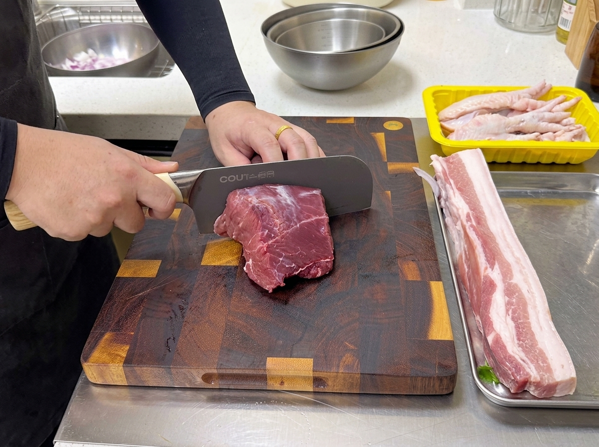 木製のまな板の上で、大きな生の牛肩ロース肉の塊をプロ用の包丁で2つに切り分ける様子。