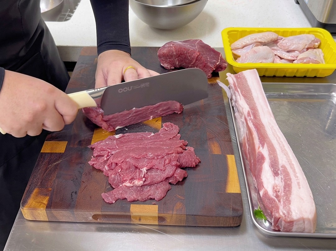 まな板の上で、包丁を使って生の牛肩ロース肉を繊維に対して垂直に薄切りにする様子。