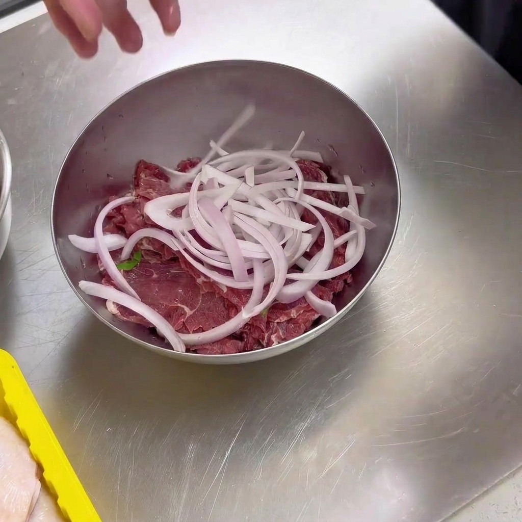 ステンレス製のボウルに入った生の薄切り牛肉の上に、スライスした新鮮な白玉ねぎをのせた状態。