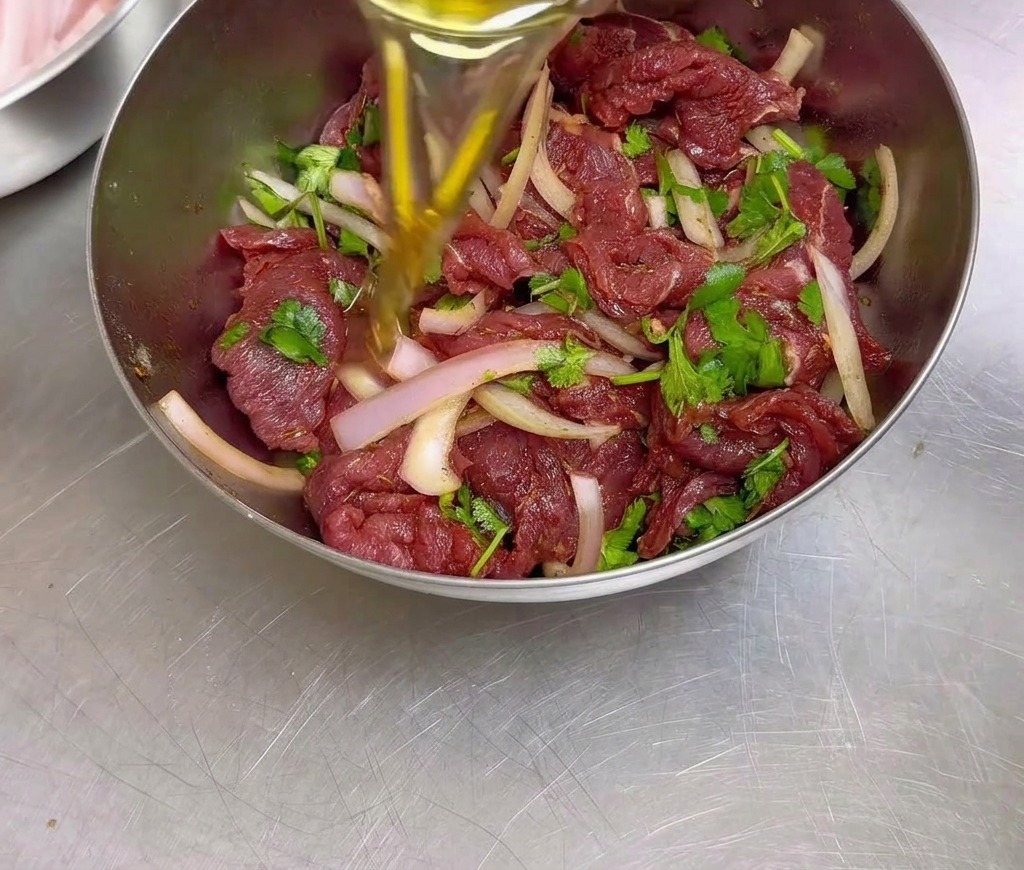 味付けが終わった牛肉、玉ねぎ、香菜が入ったステンレスボウルに、サラダ油を少しずつ注ぎ入れる様子。