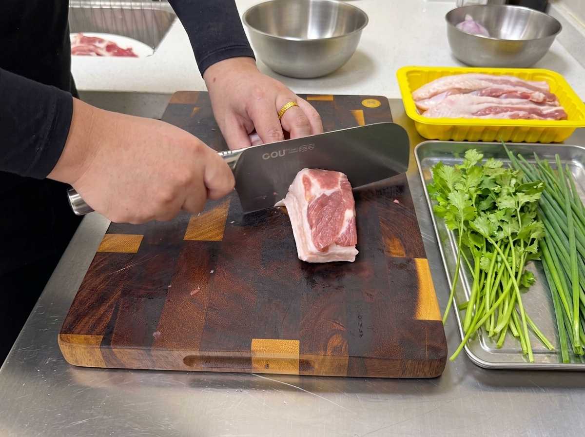 まな板の上で、脂身のバランスが良い生の豚バラ肉の塊を、大きめの包丁で均一な薄切りにカットしていく様子。