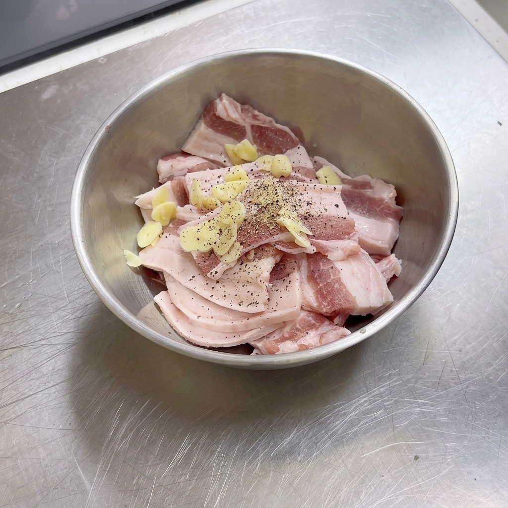 ステンレスボウルに入った生の豚バラ肉のスライスに、刻みにんにくと挽きたての黒こしょうが振りかけられている状態。