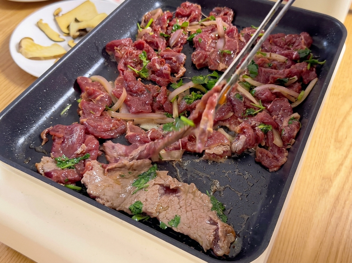 熱したホットプレートの上に、トングを使って玉ねぎや香菜と混ぜ合わせた薄切りの漬け込み牛肉を並べていく様子。