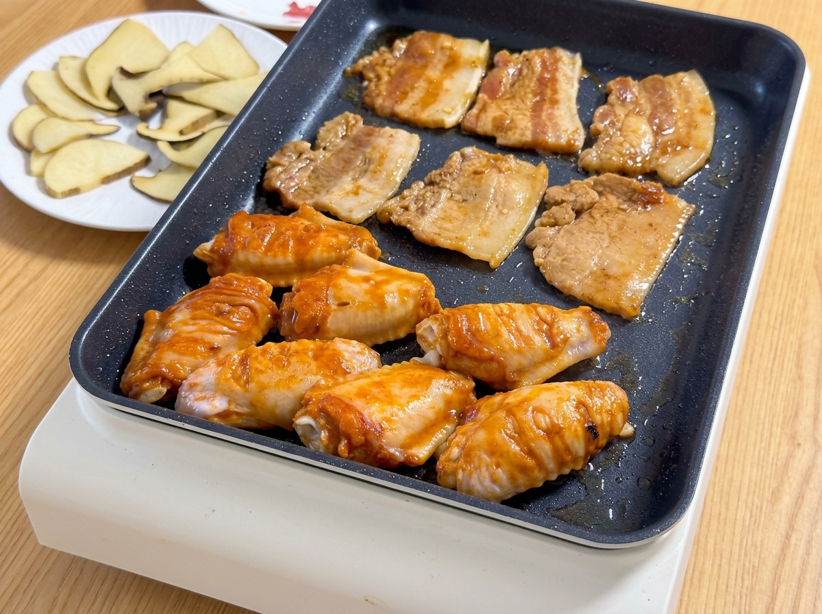 熱い卓上ホットプレートの上で、味付けされた豚バラ肉のスライスと、鮮やかなオレンジ色のオルレアン風手羽先が一緒にジューシーに音を立てて焼けている様子。