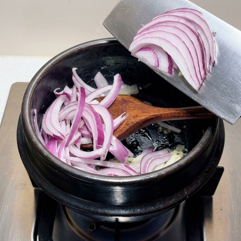 香味野菜が入った黒い調理鍋に、幅広の包丁の刃からスライスした紫色の赤玉ねぎを移し入れている様子。