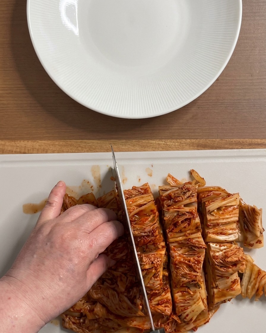 白いまな板の上で、鮮やかな赤色の大きな熟成キムチの塊を食べやすい大きさに切り分ける手元。