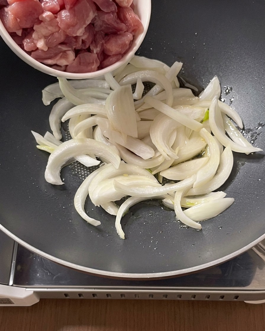 スライスした玉ねぎとオイルが入った鍋に、小さなボウルから生の豚肉の切れ端を加えている様子。