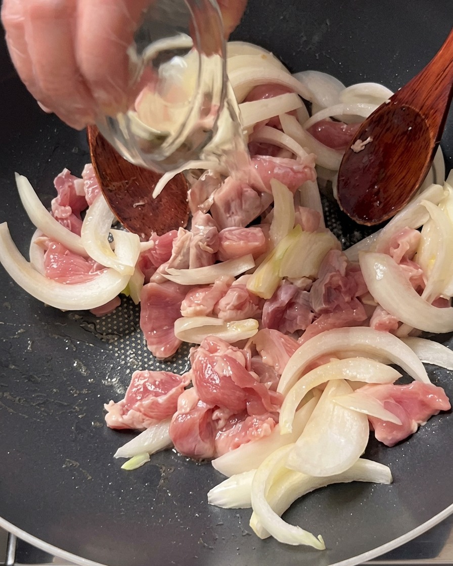 鍋の中の豚肉と玉ねぎの上へ、小さなガラスのボウルから料理酒を注ぎ入れている様子。