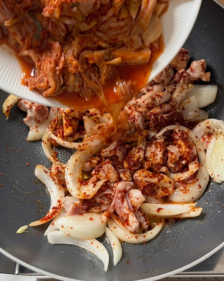 下味をつけた豚肉と玉ねぎが入った鍋に、白いお皿から刻んだキムチを投入している様子。