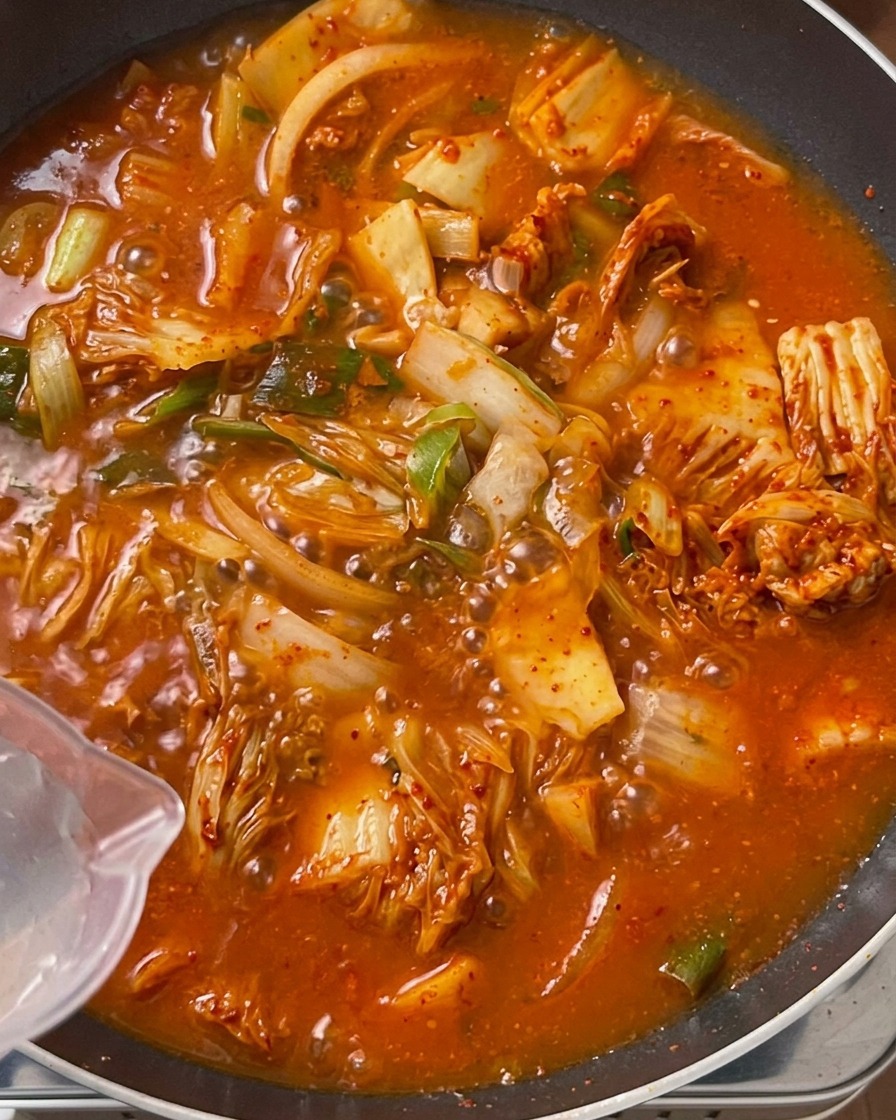 炒められて鮮やかなオレンジ赤色になったキムチとお肉の入った大きな鍋に、透明なプラスチックのピッチャーからきれいな水を注ぎ入れている様子。