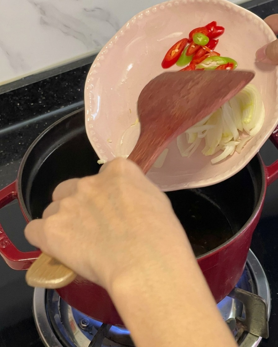 木べらを使って、小さなピンクの皿から刻んだ赤・青唐辛子と香味野菜を、油のひかれた赤い鍋に移している様子。