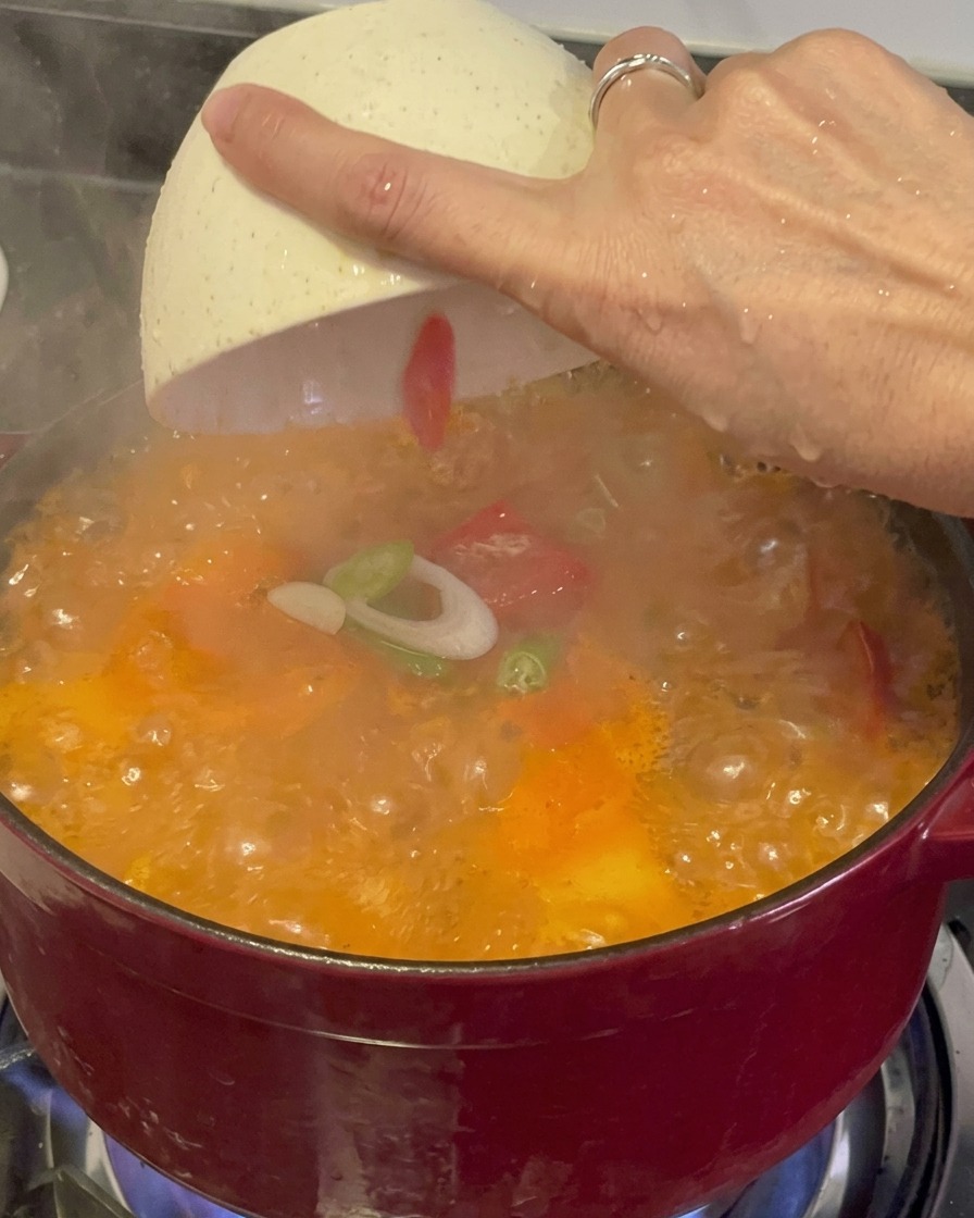 湯気が立ち上りグツグツと煮える赤いキムチチゲの上に、白いボウルから刻んだ青ねぎと赤唐辛子を散らしている様子。