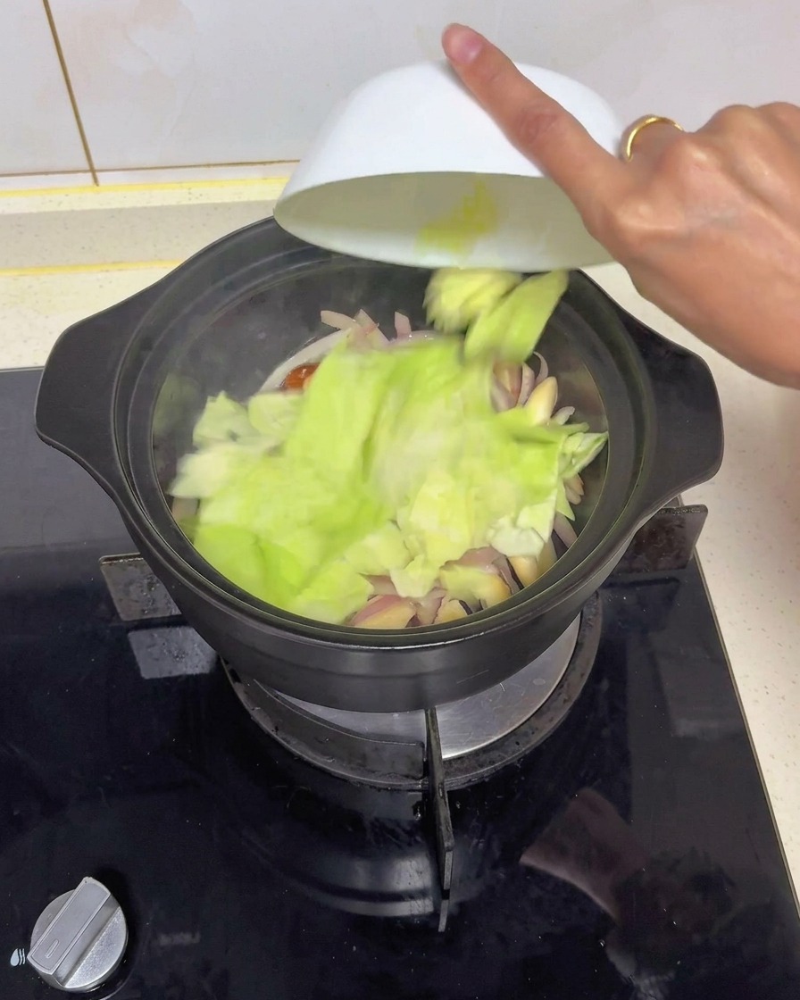 炒めた玉ねぎと椎茸が入った黒い鍋に、白いボウルからざく切りにしたキャベツを投入する様子。