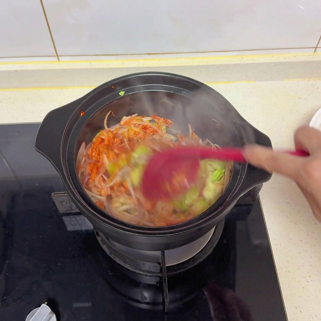黒い鍋の中で、赤いスパチュラを使って野菜とキムチを炒めながら、醤油や塩を馴染ませる様子。