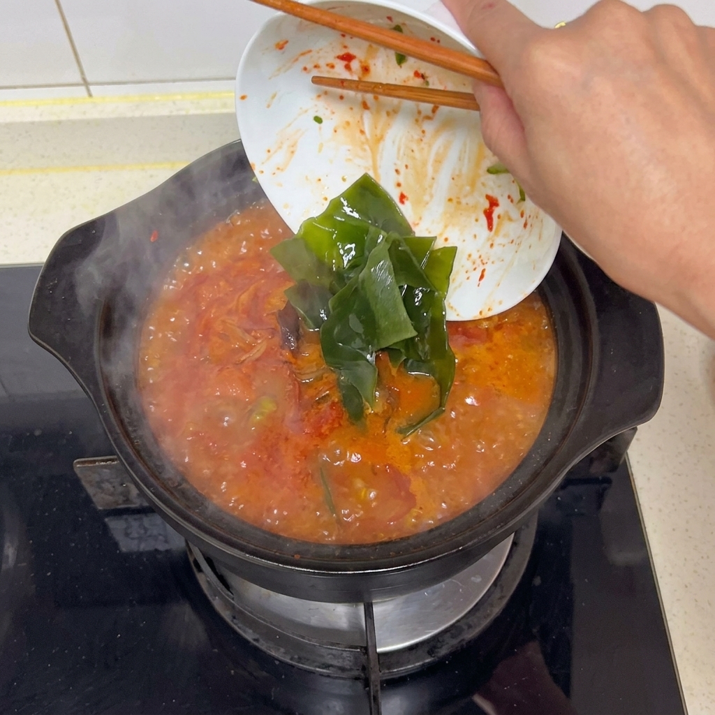 グツグツと沸騰する真っ赤なスープに、箸を使って白い皿から戻した緑色のだし昆布を入れる様子。