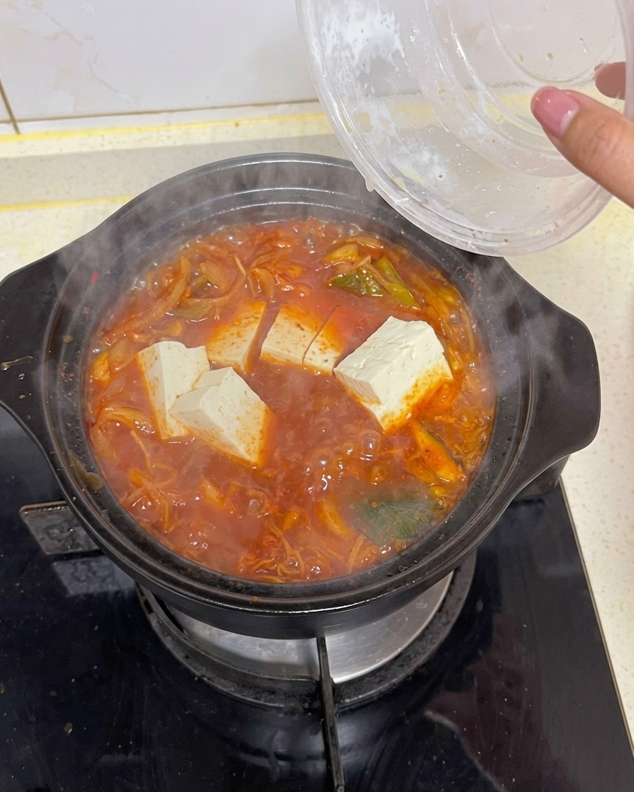 青菜や昆布が入った真っ赤なキムチスープに、大きな白い長方形の豆腐を投入し、グツグツと煮立てている様子。
