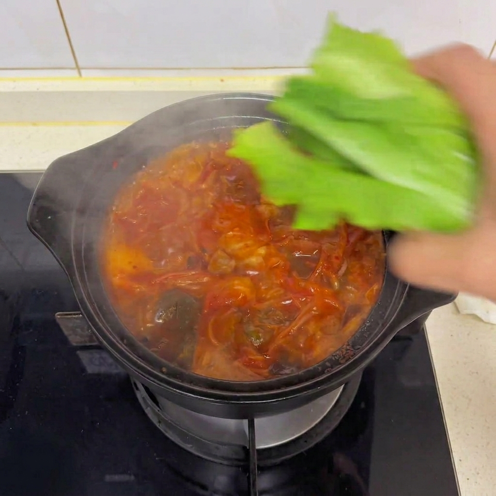 湯気が上がる熱々のキムチ豆腐スープの上に、新鮮な緑のロメインレタスを手でちぎって乗せる様子。