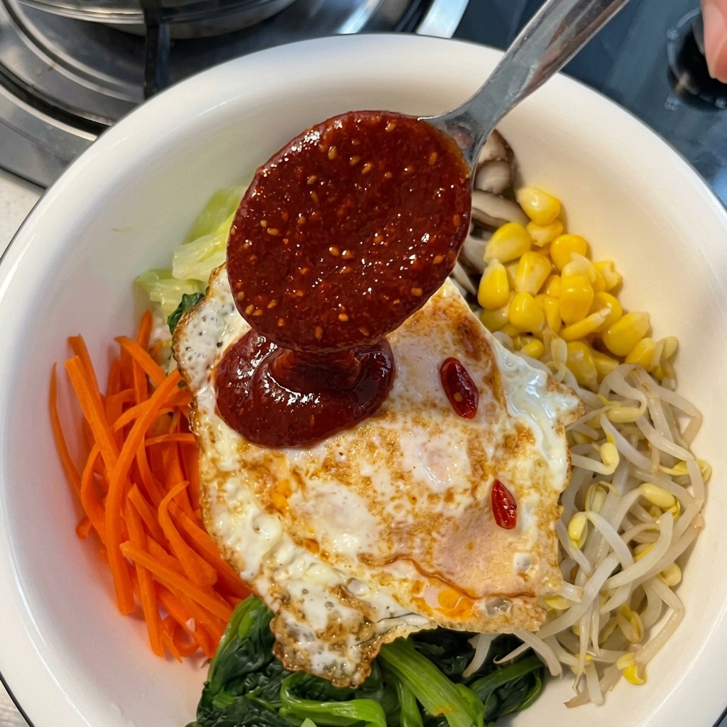 カラフルなナムルと目玉焼きがのったビビンバ丼に、金属製のスプーンから濃厚な赤色のコチュジャンソースを回しかけている様子。