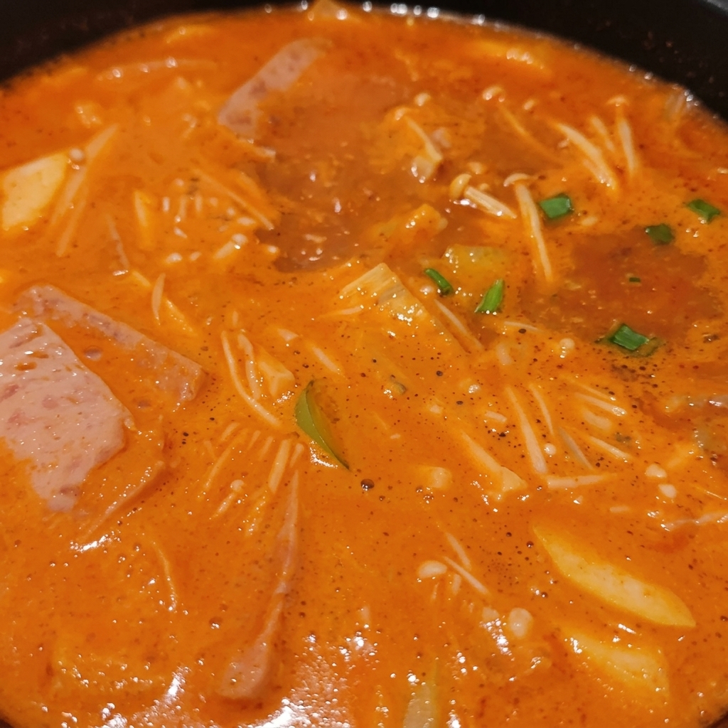 エノキダケ、肉の薄切り、刻みネギ、豆腐が濃厚なオレンジ色のスープに浸かり、ぐつぐつと煮え立っている韓国チゲ。