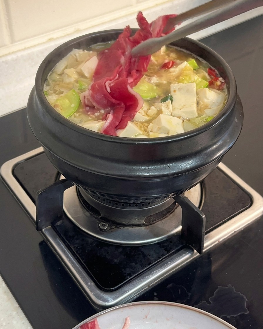料理用トングを使い、豆腐やズッキーニが入って沸騰している韓国風チゲの鍋へ、生の牛薄切り肉を投入している様子