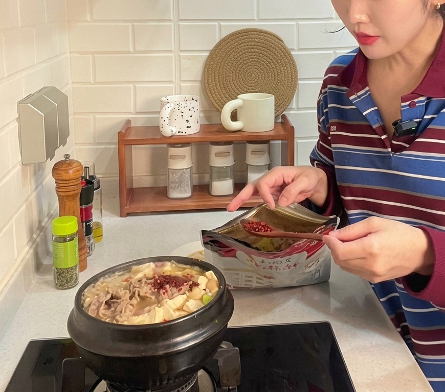 豆腐や薄切り肉がたっぷり入ってグツグツと沸き立つ土鍋のテンジャンチゲに、スプーンで粗挽きの赤粉唐辛子が加えられている様子
