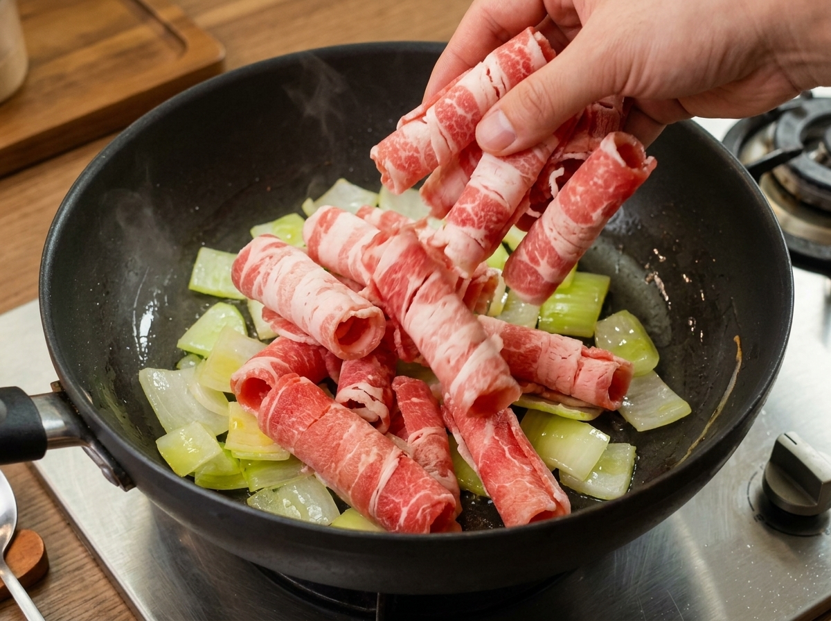 しんなりと炒めた半透明の玉ねぎが入った鍋に、生の牛薄切り肉を加えている様子。