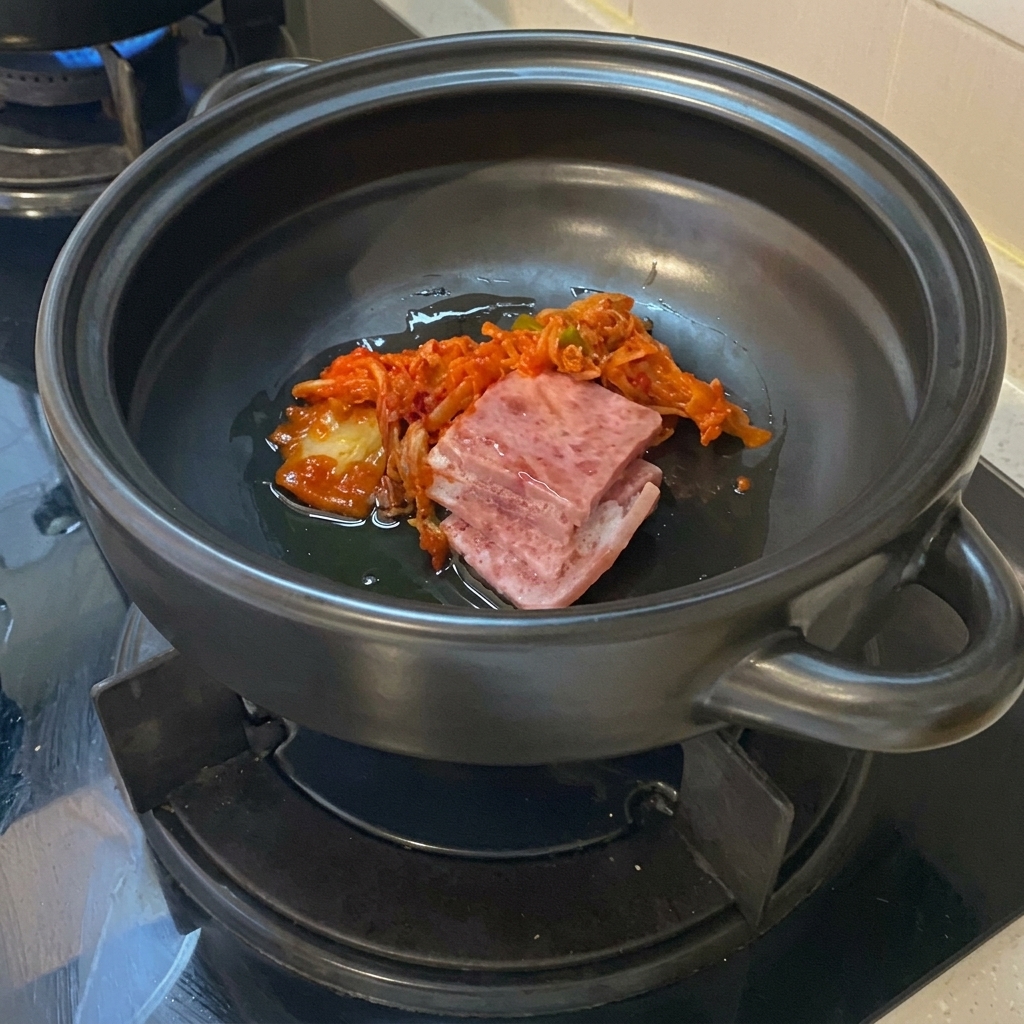 黒い土鍋の底で、油と一緒に炒められている生の豚バラ肉のスライスと鮮やかなキムチ。