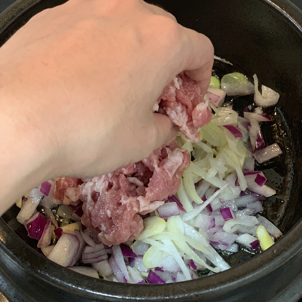 長ねぎと玉ねぎを炒めた鍋に、一口大の生豚バラ肉を加えている様子。