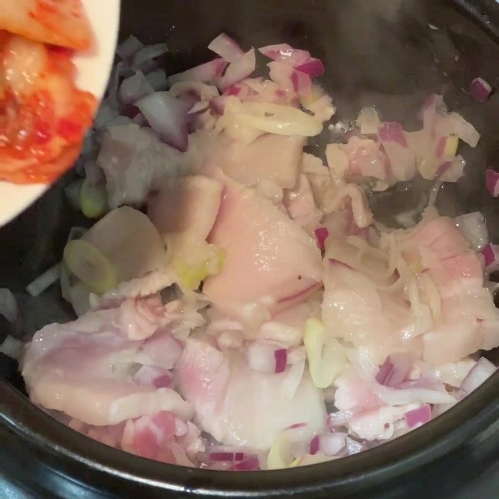 豚バラ肉と玉ねぎが入った鍋に、お皿から刻んだキムチを加えている様子。