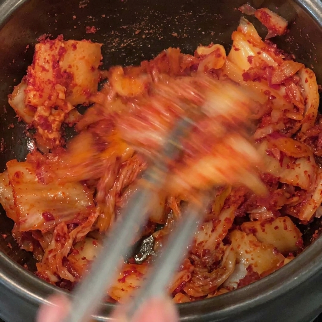 黒い鍋の中で、赤い粉唐辛子をキムチや豚肉全体にしっかりと混ぜ合わせながら炒めている様子。