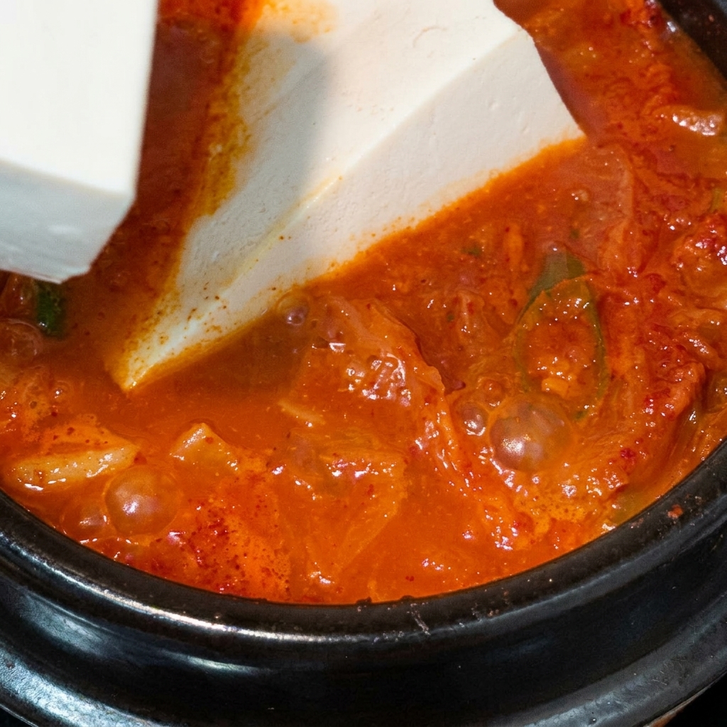 グツグツ煮立つ赤いキムチスープの中に、大きな絹ごし豆腐の塊を慎重に入れている様子。