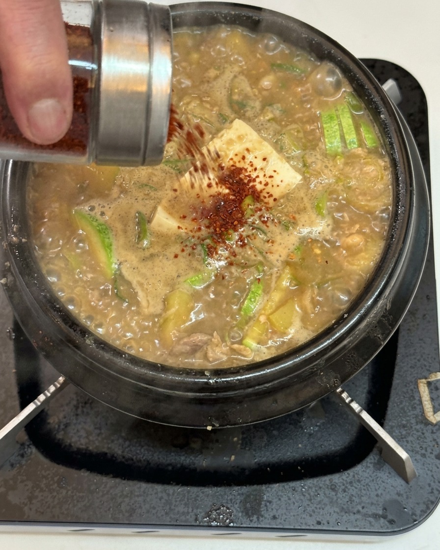 豆腐やズッキーニが入ってぐつぐつと煮えるチゲの上に、ガラス瓶から赤い粉唐辛子を振りかけている様子。