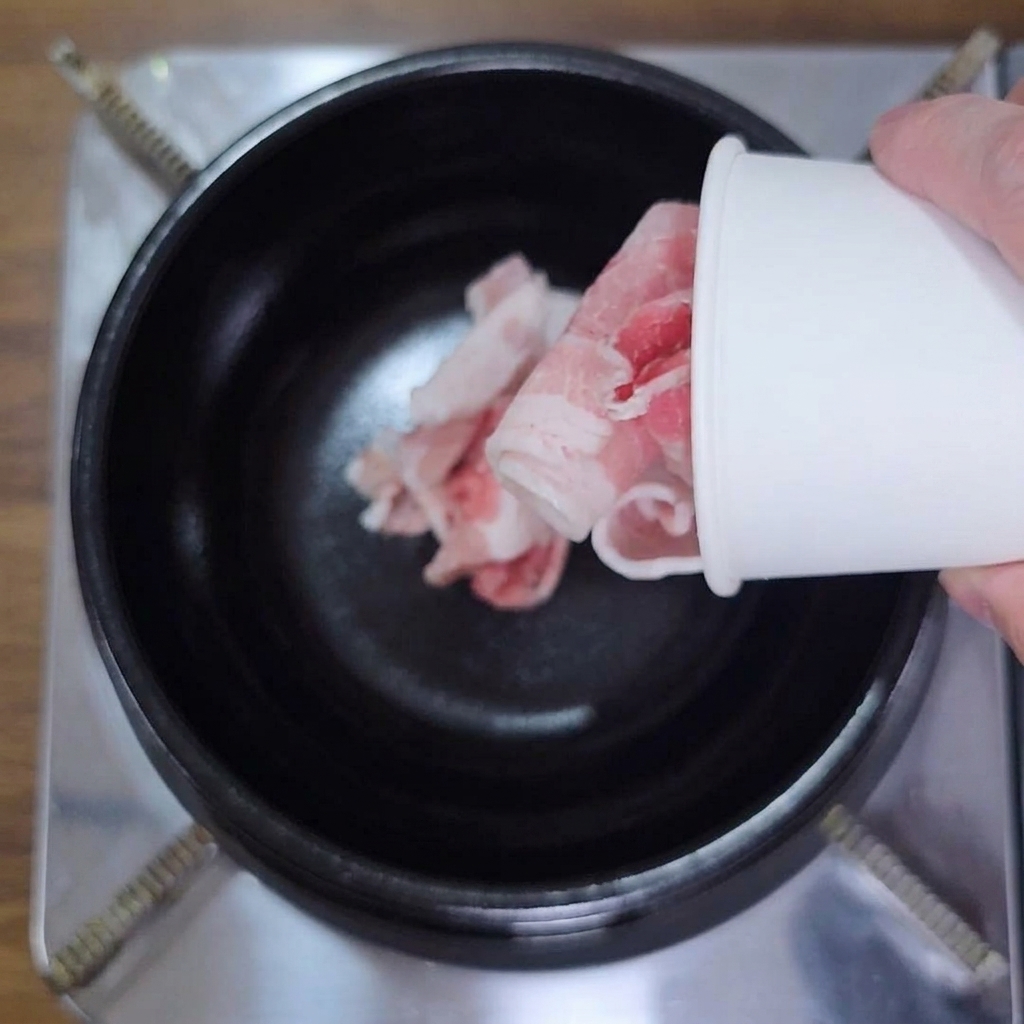 丸まった薄切りの生豚バラ肉を紙コップから黒い鍋に入れる様子。