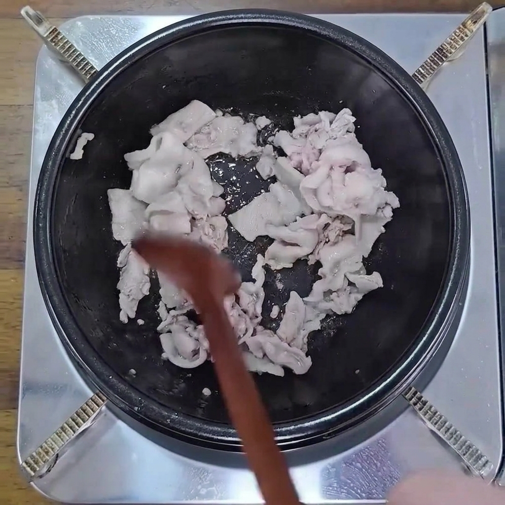 木べらを使って黒い鍋で薄切りの豚バラ肉を軽く焼き色がつくまで炒めている様子。