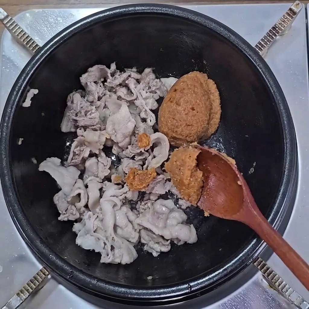茶色のテンジャンとサムジャンを鍋の中の炒めた豚肉の上に加える様子。