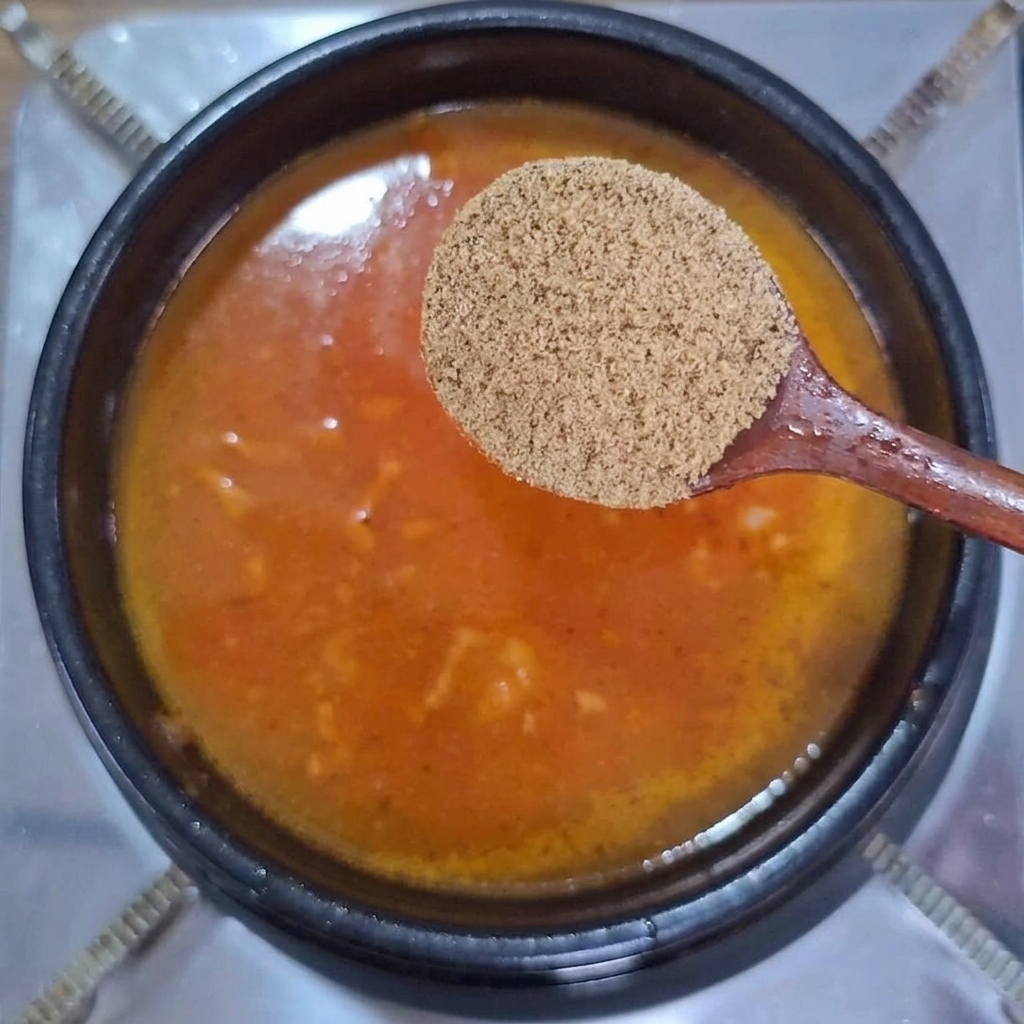 沸騰する赤オレンジ色のスープの上で、木べらに乗った牛だし粉末を持っている様子。
