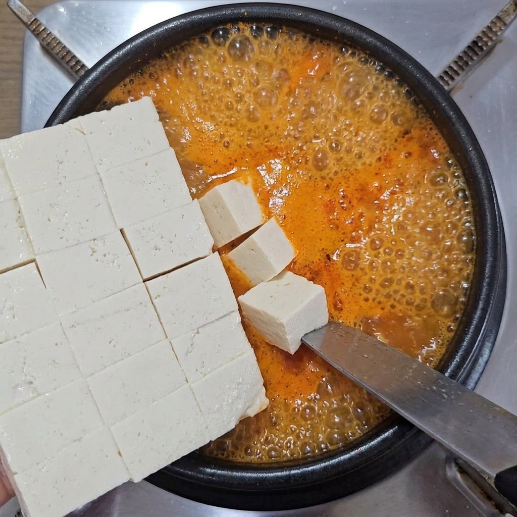 大きなナイフで角切りにした白い豆腐を、沸騰する鍋の中にそっと滑り込ませている様子。