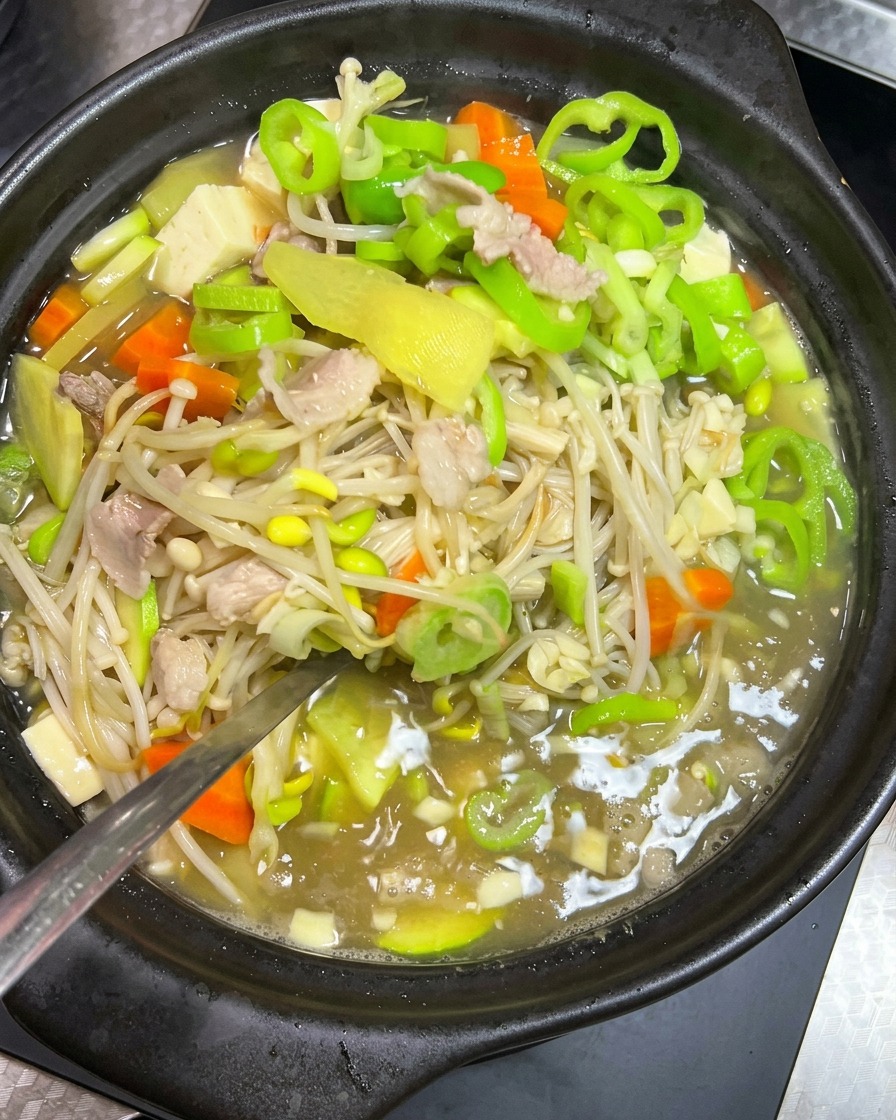 黒い土鍋の中で煮立っている豆もやし、豚肉、緑の野菜のスープを、金属製のお玉で優しくかき混ぜている様子。