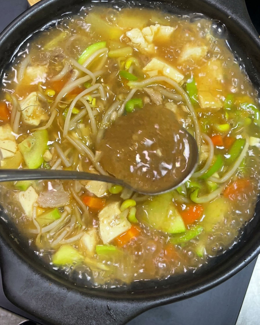 コチュジャンを乗せた金属製のお玉を、豆腐やズッキーニ、豆もやしが入った沸騰する土鍋のスープに入れて溶かしている様子。