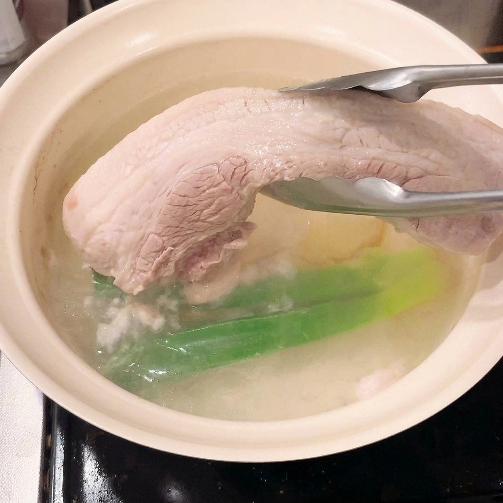 トングで下茹でされた豚バラ肉を鍋から引き上げているところ。