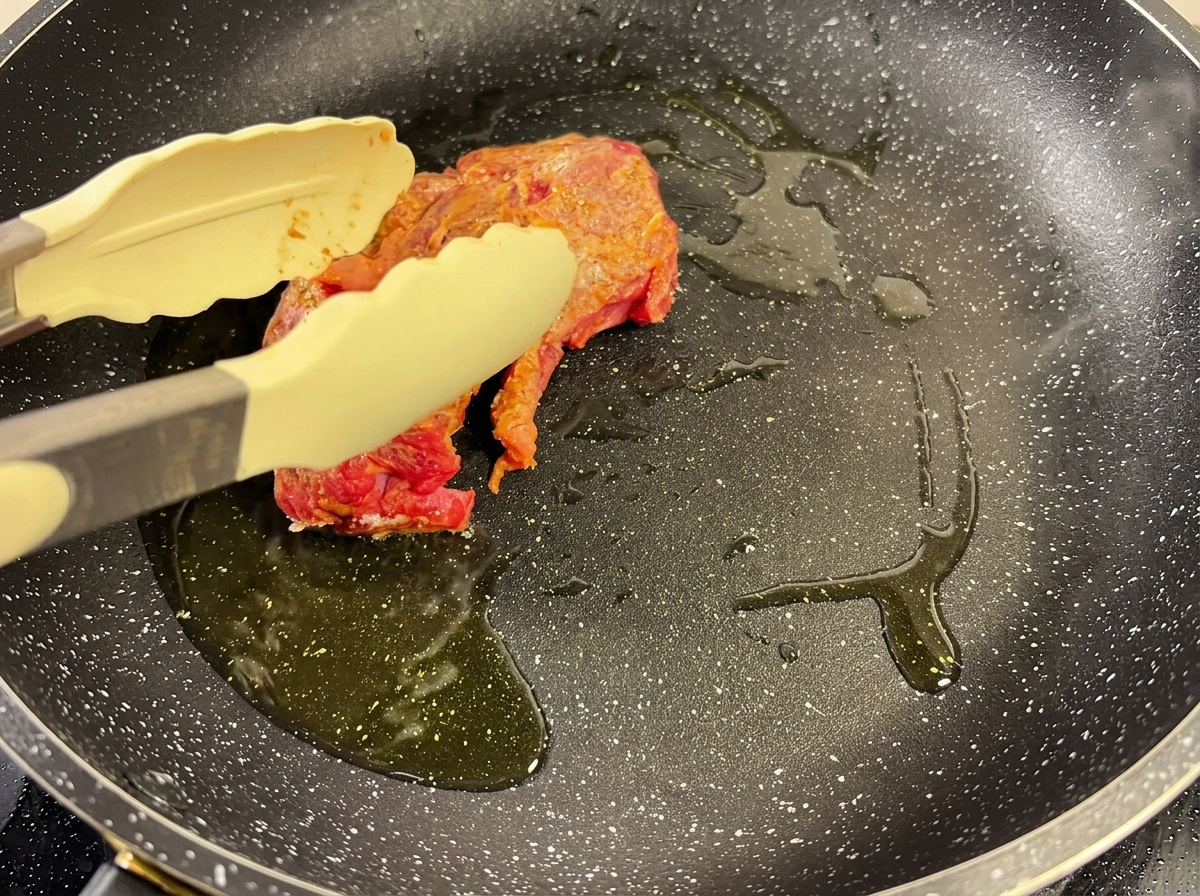 熱したフライパンに油を引き、トングで押さえながら味付けした牛肉の表面を焼き付けている様子。